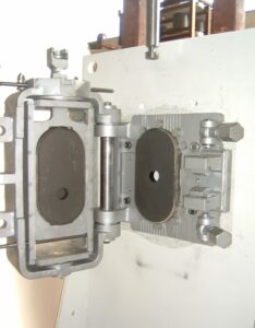 Das Bild zeigt einen FLO-CON Schieber der Firma Knöllinger FLO-TEC, Typ 6317.3 ohne Schutzplatte, der an einem Montagebock montiert ist. Der Dreiplatten-Gießpfannenschieber ist im aufgeklapptem Zustand gezeigt und die feuerfesten Platten sind zu sehen. Die beiden Schwenkschrauben, mit denen der Schieber verriegelt wird sind beiseite geschwenkt.